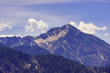 A scenic view of the Ringspitz mountain rising behind the lakeside town of Bad Wiessee in Bavaria, framed by forested slopes and a clear alpine sky.
