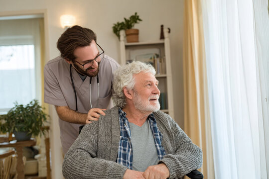 Male nurse providing help to elderly man who is disabled, sitting in wheelchair, giving assistance to disabled old people. Senior pensioner talking to physiotherapist being cared in nursery house