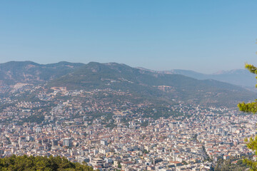 Fototapeta premium Vibrant urban development fills the foreground with an expansive city layout. Majestic mountains rise in the background under a clear blue sky