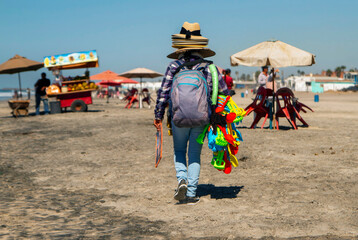 A vendor carrying hats and sand toys walking along a beach. A vendor with a cart is in the background