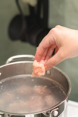 Woman's hand dropping frozen chicken meatball into boiling pot for homemade soup preparation, cooking process close-up with steam