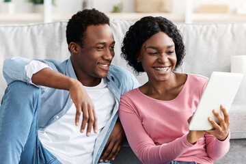 Cheerful black couple with cleaning tools resting, sitting on floor in living room, using digital tablet pad, looking for house-keeping service on Internet