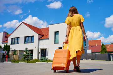 Woman in yellow dress pulls orange suitcase along residential street with houses on sunny day. Female traveler returning home from journey. Concept of travel, homecoming and end of vacation