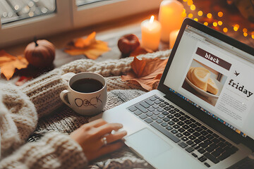 Cozy autumn setting with woman shopping online for Black Friday deals on laptop while drinking coffee, surrounded by candles, pumpkins and warm blanket by the window