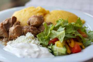 A plate of delicious traditional Moldovan food featuring yellow hominy, tender meat, fresh salad with cucumbers and tomatoes
