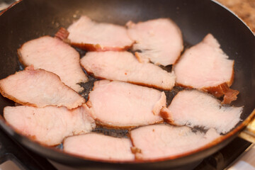 Slices of meat cooking in a frying pan. Meat is light pink with a crispy edge, arranged evenly in pan