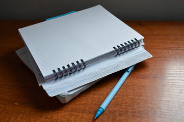 a stack of squared notebooks with a blue pen lying on a wooden desk, top notebook is spiral-bound...