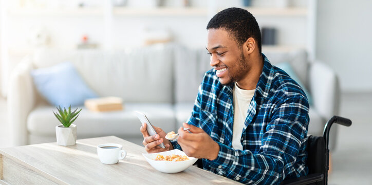Happy black guy in wheelchair eating breakfast and browsing web on smartphone at home, copy space. African American man posting in social media or watching video online during his meal - Powered by Adobe