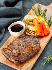 Fresh hot pork steak on wooden cutting board, served vegetables and sauce. Delicious pork steak on dark table. BBQ rib beefsteak or tender pork steak