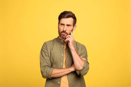 A middle aged man in his 40s stands against a vibrant yellow backdrop. He has a beard and casual attire, resting his hand on his chin, deep in thought and contemplation.