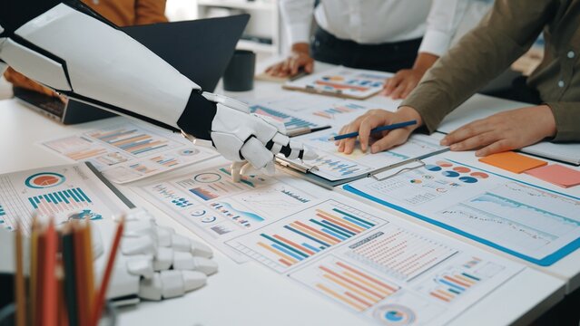 A robotic arm aids human colleagues in a modern office environment, focusing on data analysis and strategic planning with various reports and charts displayed.Noogenesis