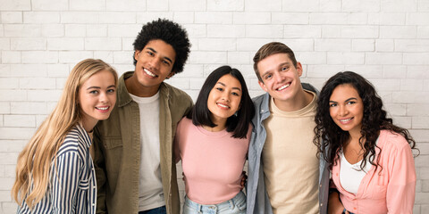 Friendship of exchange students and modern education. Happy young multiracial people team in casual hugging in college or university, smiling and looking at camera, on white brick wall background