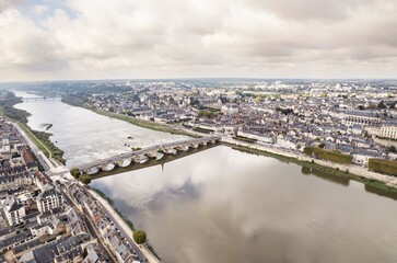 An expansive river divides a cityscape under a heavy cloud cover. A stone bridge connects both sides of the water, leading to a dense area of buildings and green spaces