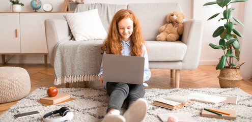 Modern Lifestyle. Smiling young teenage girl with ginger hair sitting on the floor carpet, leaning on couch, using laptop and typing on keyboard. Schoolgirl chatting with friends online on internet