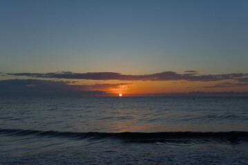 The sun rises on the horizon over a calm, dark ocean, painting the sky with warm orange, pink, and purple hues and scattering light across the water.