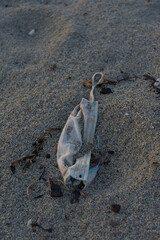 A dirty, crumpled white disposable face mask lies on a sandy beach, surrounded by small pieces of dark debris.
