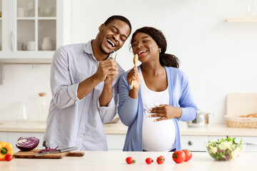 Positive pregnant black couple singing while cooking at home, happy african american expecting lady making dinner with her husband, singing songs and dancing, kitchen interior. Family, love concept
