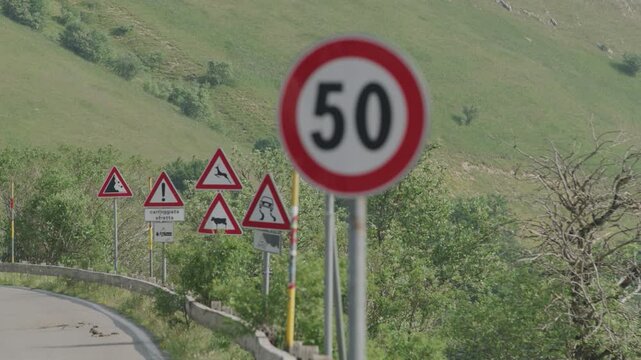 Speed Limit Sign on Rural Road with Warning Signs and Lush Green Hills
