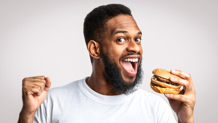 Joyful Black Guy Biting Burger Enjoying Cheat Meal Eating Unhealthy Cheeseburger Standing Posing...