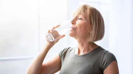 Fit senior woman drinking clear water during her workout break at home. Mature Caucasian lady staying hydrated after sports training. Healthy lifestyle and wellness concept