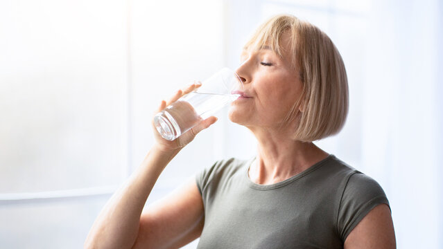 Fit senior woman drinking clear water during her workout break at home. Mature Caucasian lady staying hydrated after sports training. Healthy lifestyle and wellness concept