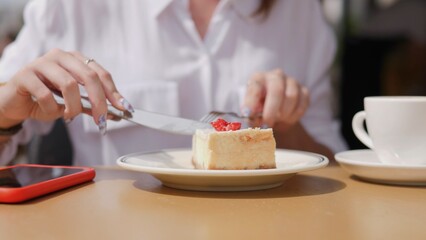 A woman sits at a table, cutting a slice of cake with a fork and knife, enjoying coffee in a bright outdoor setting during summer. Friends connect and share laughter.