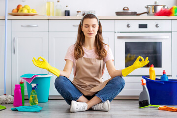 Young housewife surrounded by cleaning supplies meditating on floor at kitchen, taking break from house chores. Millennial maid resting from domestic duties, finding balance between life and home work