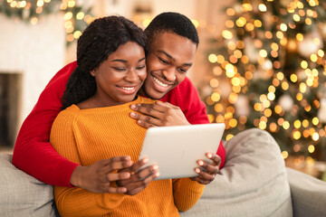 Video call on eve of Christmas. Family happy millennial african american husband and wife look at tablet and congratulate friends in cozy living room interior with Xmas tree and glowing garlands