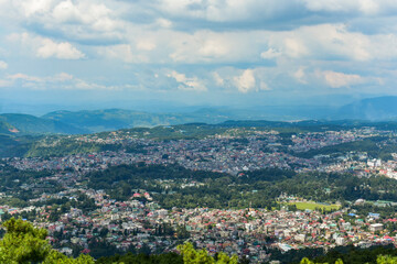 Shillong city nestled amidst hills and lush greenery, showcasing a panoramic view under a cloudy sky.