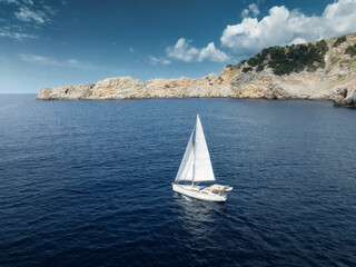 Aerial drone view of a sailboat sailing on blue Mediterranean sea near rocky coastline and cliffs...
