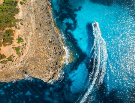 Aerial drone top view of motorboat making waves near rocky coastline and turquoise sea in bright sunlight. Mallorca, Balearic Islands, Spain. Mediterranean summer landscape, travel and adventure. Boat