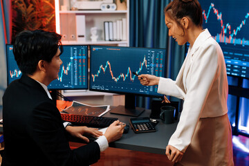 Smiling Asian business traders pointing to dynamic stock exchange data screen profit value online website program at night time at home office. Analyzing currency rate value on data report. Infobahn.