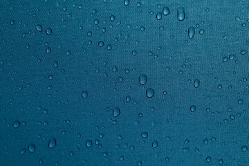 Macro photograph of water droplets on teal blue waterproof fabric. The close-up reveals the texture of the material and the natural round shapes of condensation, creating a clean and minimalist backgr
