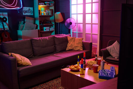 Living room scene showing empty gray sofa with snacks, drinks, foam finger and party supplies on coffee table, illuminated by colorful neon and ambient lighting, bookshelf and guitar in background