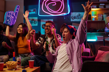 Group of young adults including Asian woman, Black man, Caucasian man cheering and celebrating while watching sports event on television, raising hands and holding drinks in living room