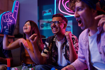 Group of young adult multiethnic friends cheering and celebrating while watching sports event on television, showing excitement and enthusiasm with raised hands and foam finger