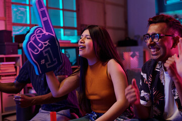 Young adult Hispanic woman cheering with oversized foam finger while sitting next to young adult South Asian man smiling, both watching sports event indoors with excitement