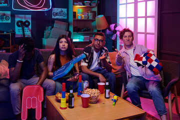 Group of four young adults, including Black man, Asian woman, South Asian man, and Caucasian man, sitting on couch watching television, holding snacks and drinks, showing mixed emotions