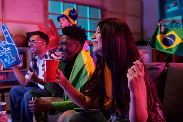 Group of young adult multiethnic friends cheering and celebrating while watching sports event on television, holding drinks and waving Brazil flags, sitting together on couch