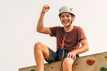 High-resolution photo of a joyful young climber sitting atop a climbing wall, raising a fist in triumph. Wearing a safety helmet and harness, the smiling teen celebrates achievement, self-confidence