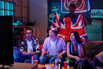 Obraz premium Group of young adults and Black man supporting British sports team, holding United Kingdom flags and watching television together, snacks and drinks on table in living room