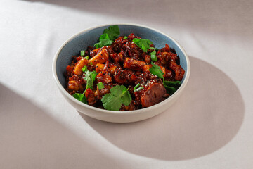Korean rice cakes with sliced sausage, mushrooms, spicy gochujang sauce, sesame seeds and greens served in ceramic bowl with natural light