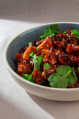 Korean rice cakes with sliced sausage, mushrooms, spicy gochujang sauce, sesame seeds and greens served in ceramic bowl with natural light