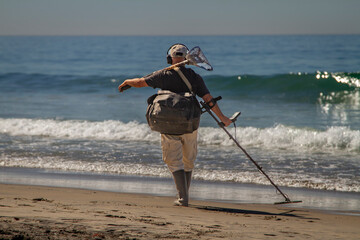 A man using a metal detector on a beach with the ocean and waves in the background