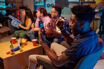 Fototapeta premium Young Black man celebrating while watching sports on smartphone, sitting in living room with diverse group of young adults cheering in background, snacks and drinks on table