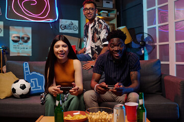 Young adult Hispanic woman and Black man sitting on sofa playing video games with controllers,...