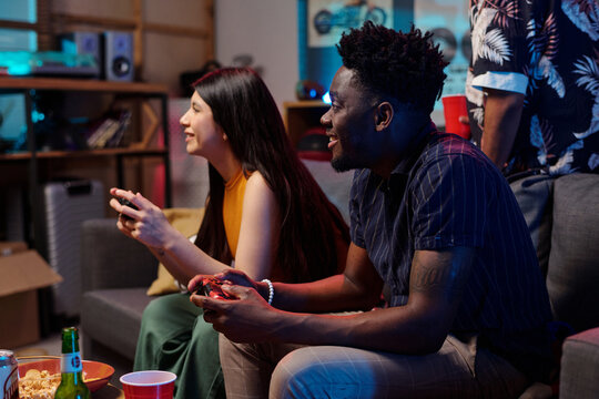 Young adult Caucasian woman and young adult Black man sitting on couch playing video game together, both holding controllers and focusing on screen, enjoying leisure activity indoors