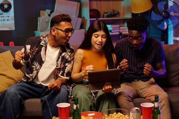 Young adult Caucasian woman sitting between young adult Caucasian man and young adult Black man using digital tablet together on couch, all cheering and reacting with excitement