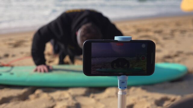 Man in hoodie exercising on surfboard on sandy beach with ocean waves, recorded on phone 