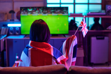 Caucasian young adult woman sitting on sofa watching soccer match on television, holding small United Kingdom flag, draping British flag over shoulders, supporting national team